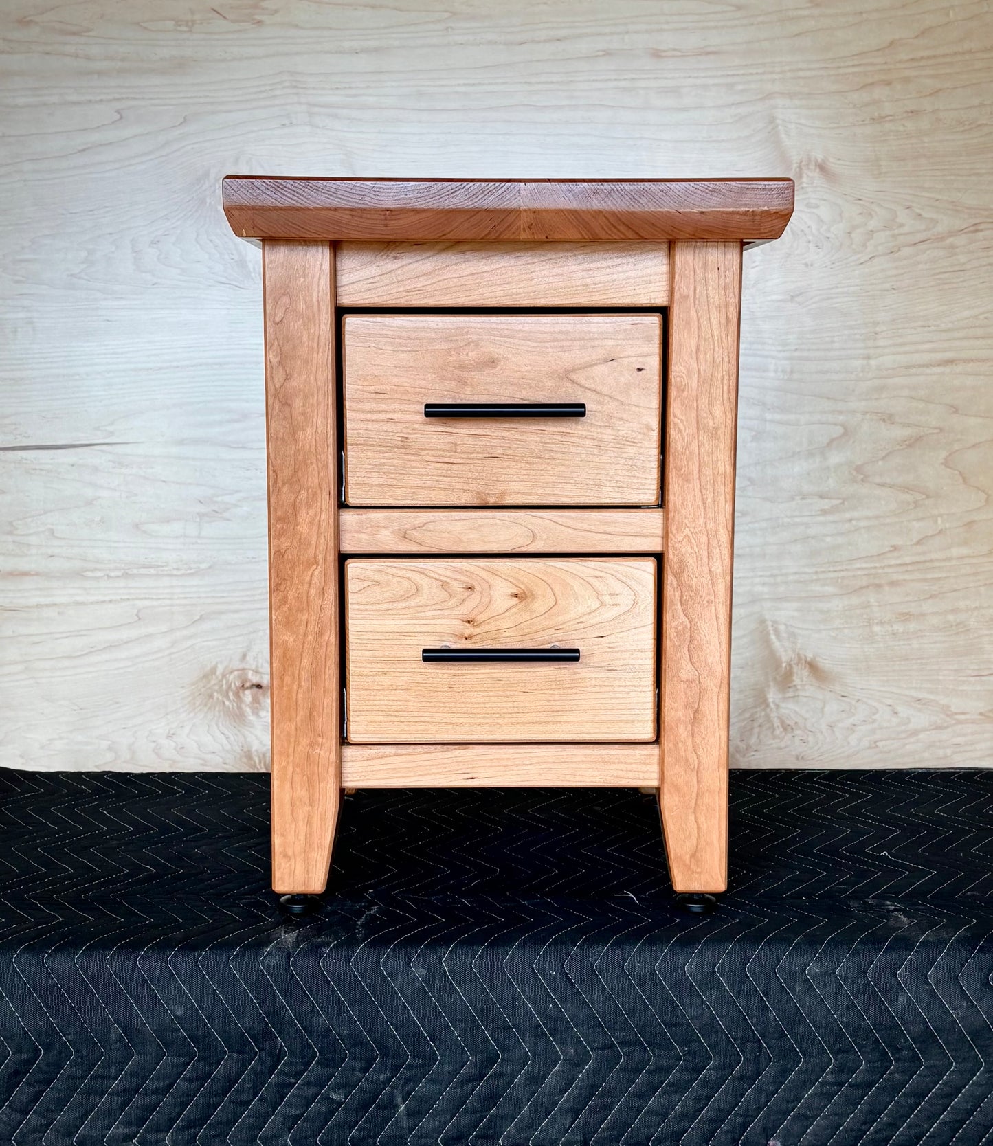Handmade Cherry, wooden nightstand with two drawers against a wooden wall.