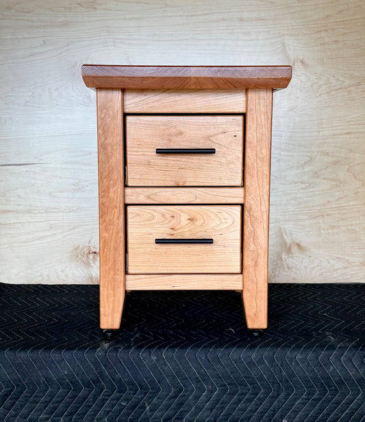Handmade Cherry, wooden nightstand with two drawers against a wooden wall.